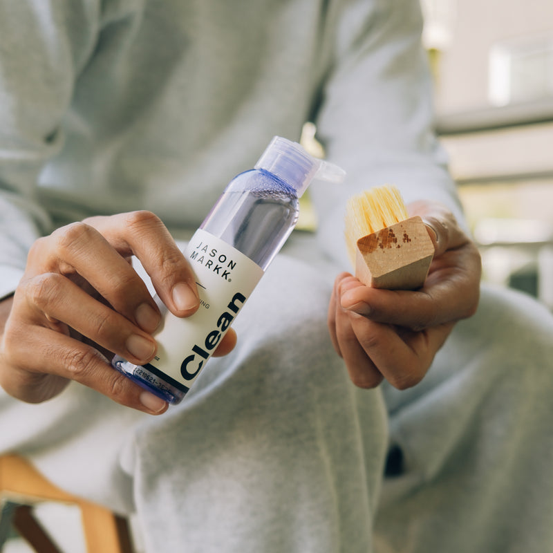 Shoe cleaner bottle with bristle brush and wooden pad held in hands.