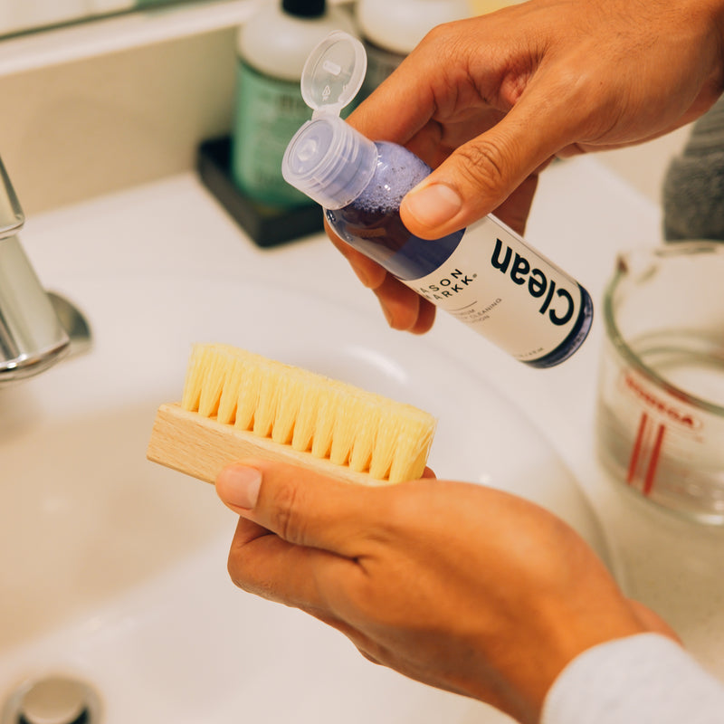 Shoe cleaner bottle held above bristle brush and foam pad.