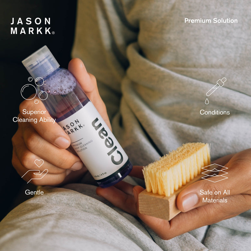 Shoe cleaner bottle in hand beside bristle brush on cloth surface.