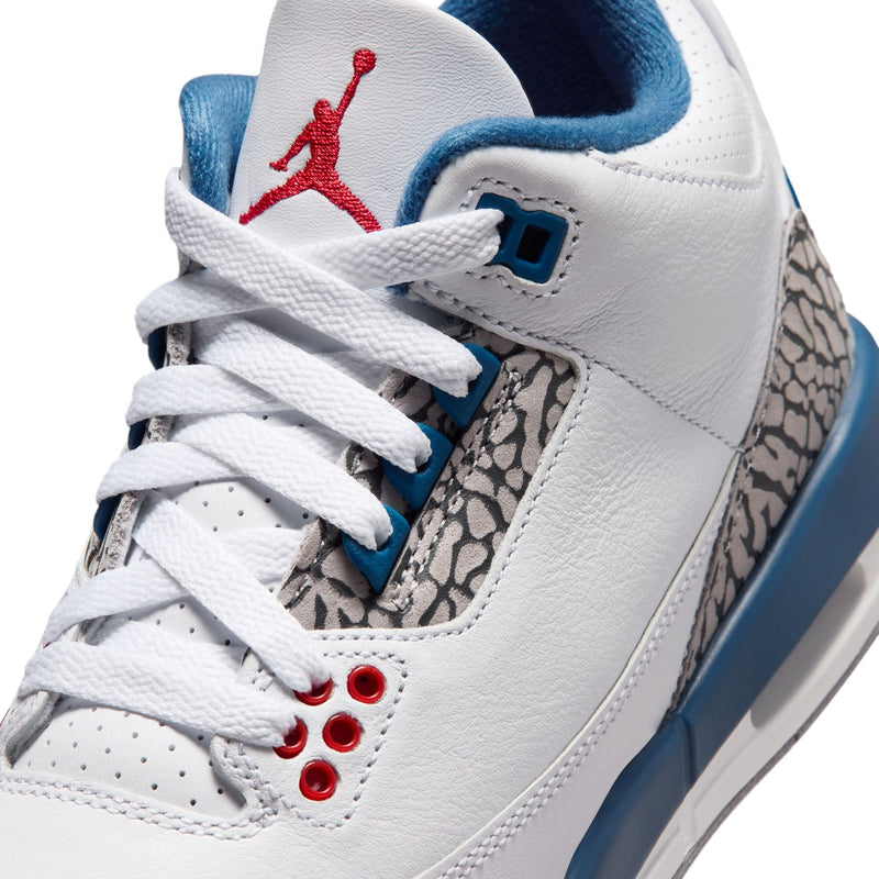 Youth white sneaker close-up with red Jumpman logo, blue lace loops, and elephant-print overlay