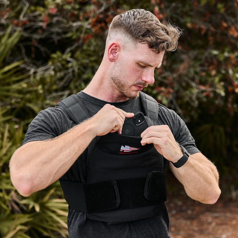 Man wearing GORUCK rucking vest outdoors, placing phone in front pocket, fitness gear