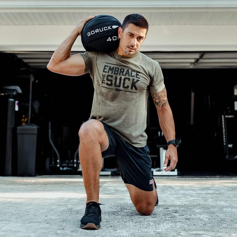 Man training with GORUCK weighted sandbag, wearing 'Embrace the Suck' shirt, outdoors