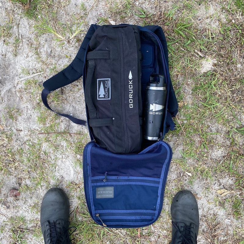 On grass, an open backpack reveals Simple Training Sandbags and a water bottle inside—ideal for sandbag workouts or your home gym. Black boots are visible below.