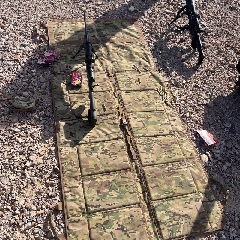A Shooting Mat is spread outdoors on rocky ground, displaying a rifle and ammunition, with another rifle partially visible.