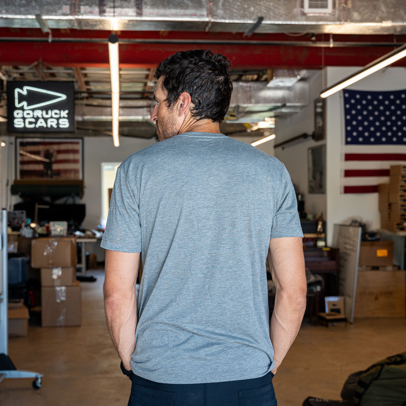 Wearing the Short Sleeve Pocket Tee - Tri-BlendX, a man stands with his back to the camera in a workshop adorned with an American flag, embodying classic simplicity and that versatile, "ruck or die" attitude.