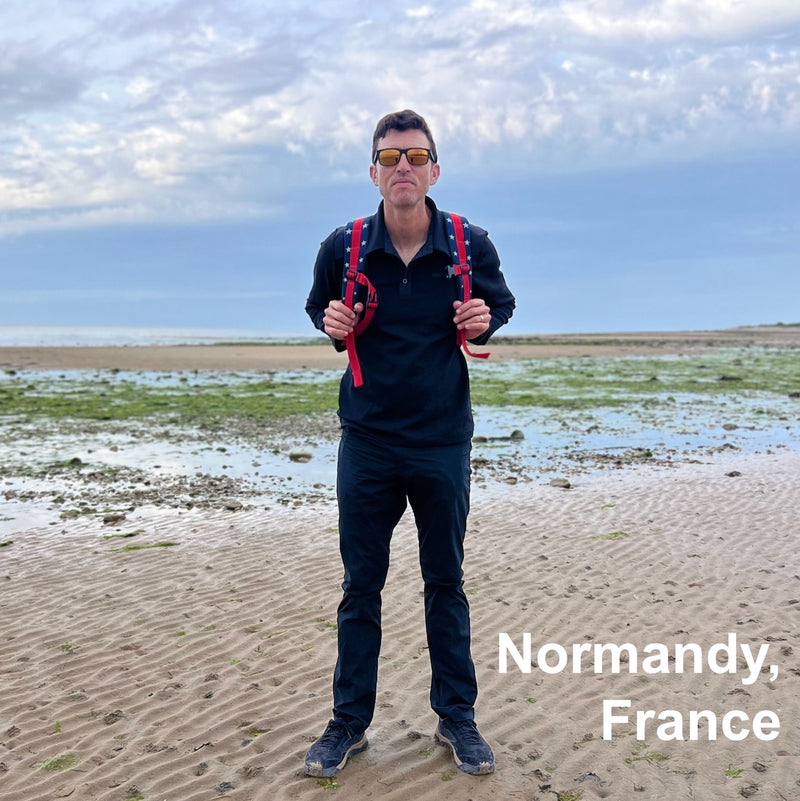 A man wearing a black outfit and red backpack stands on a sandy beach in Normandy, France, under a cloudy sky—his Size 34x32 Black only Men’s Simple Pants made from Lightweight ToughDry® fabric are ideal for anyone seeking the best pants for traveling.