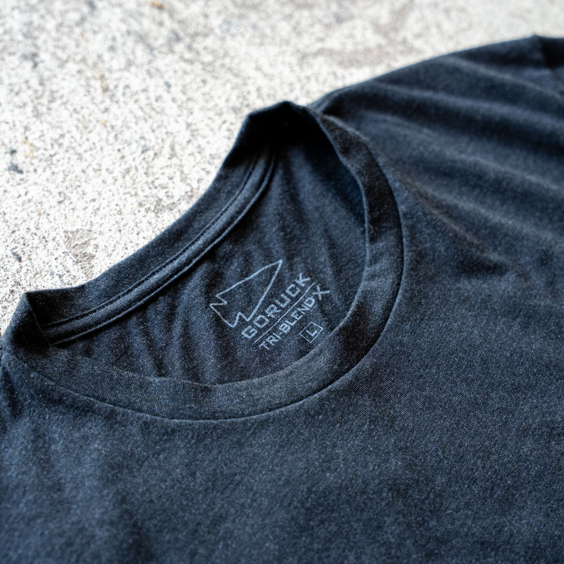 Close-up of the black Slick Short Sleeve Tee - Tri-BlendX collar with a printed label displaying the "GORUCK" logo and sizing details on concrete, showcasing its premium Tri-BlendX material for superior comfort.