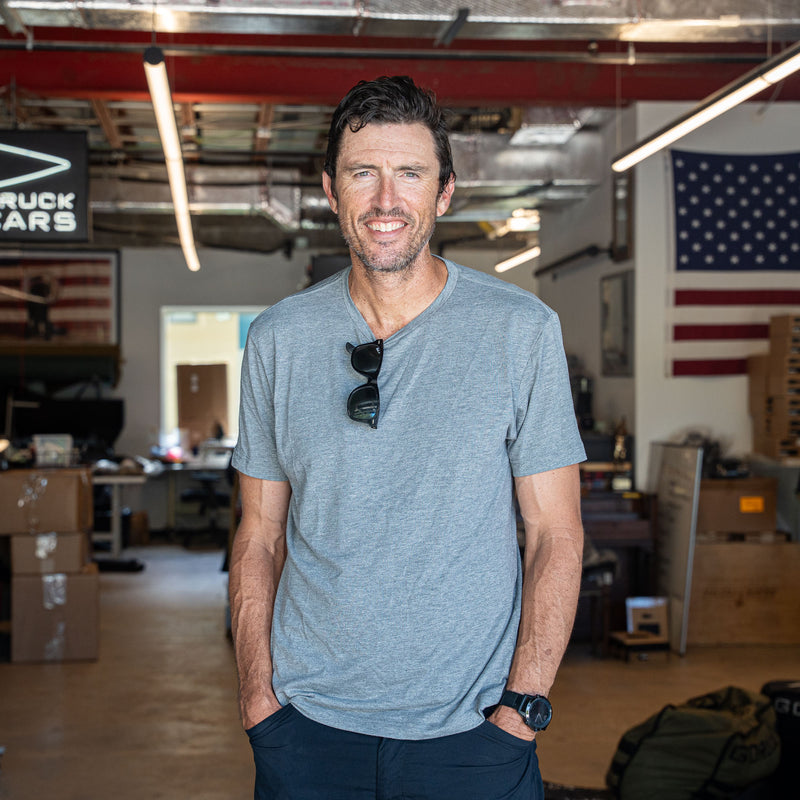 A man wearing the Slick Short Sleeve Tee - Tri-BlendX stands in a garage with American flags and boxes in the background.