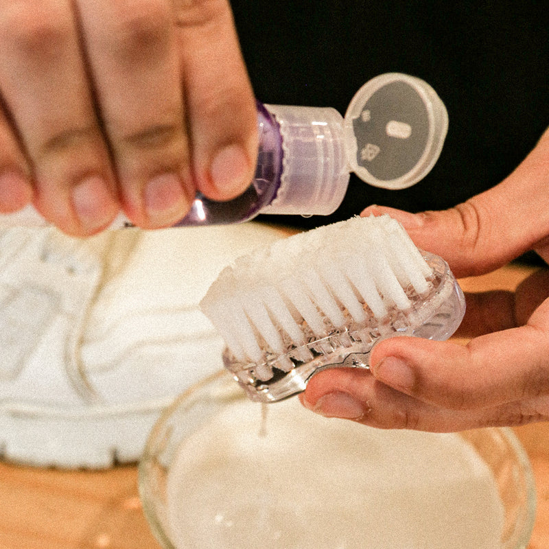Hands scrubbing a white shoe with the oval brush and deep cleaning solution