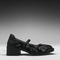 A single black leather Mary Jane-style shoe with a chunky block heel, decorative ruffles, and a strap with a metallic ring detail, shown in profile against a dark gray gradient background.