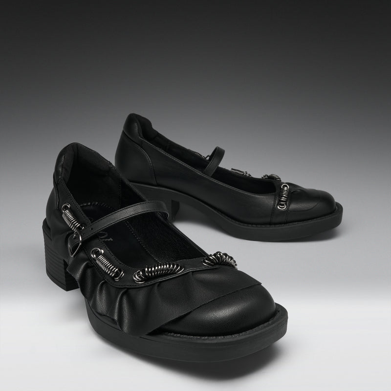 A pair of black Mary Jane-style shoes with chunky block heels, ruffled straps, and silver-tone decorative hardware, photographed on a dark-to-light gray studio background.