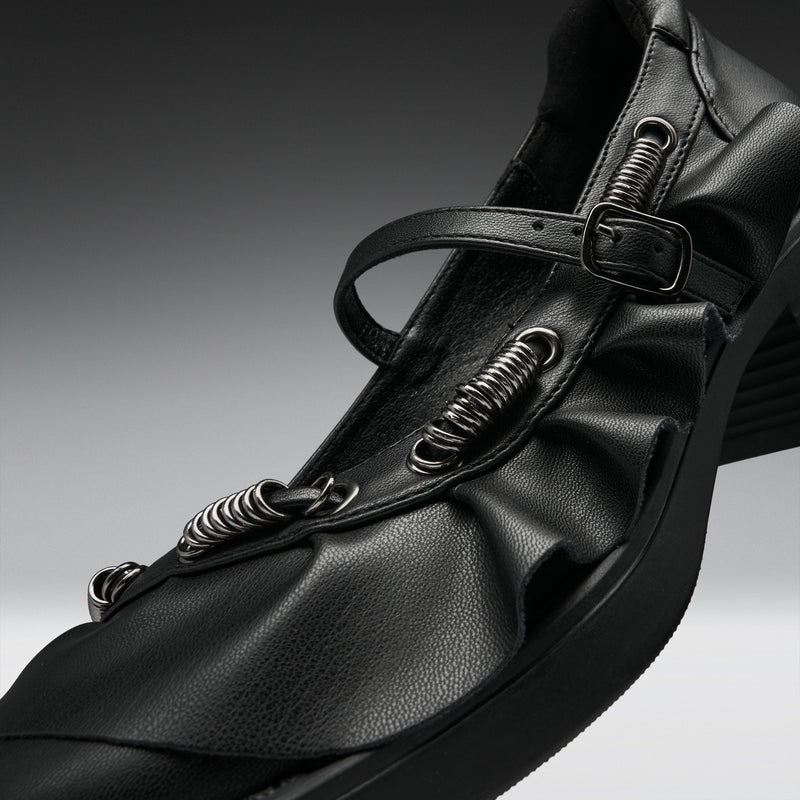 A close-up studio shot of a black leather Mary Jane-style shoe with multiple spring-loaded metal loops and a buckle strap, set against a dark gray gradient background.