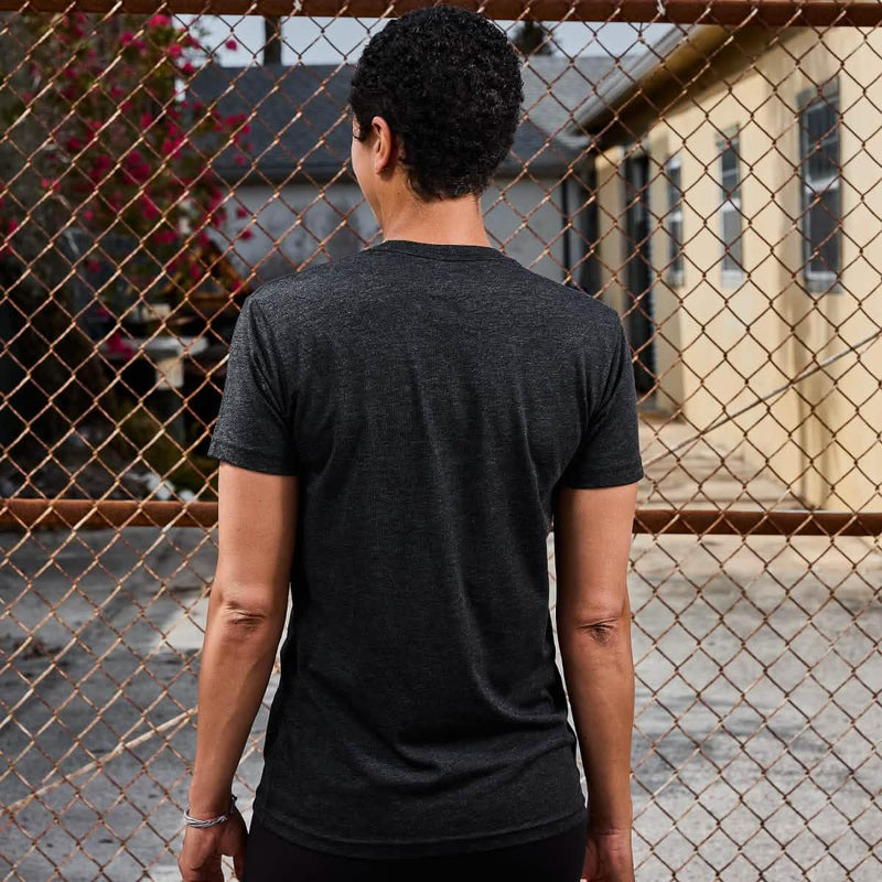 Person in dark GORUCK t-shirt standing outside by a chain-link fence, urban setting