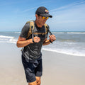 A man jogs on a sandy beach near the ocean under a clear blue sky, wearing sunglasses and a backpack with T-REX Hand Straps—his preferred rucking accessory.