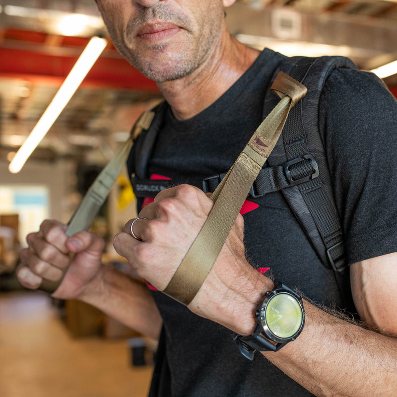 A man grips the T-REX Hand Straps, attached to his backpack straps, while wearing a black t-shirt indoors.