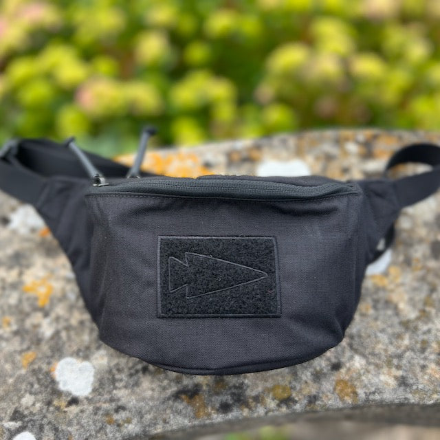 The Tactical Fanny Pack - Cordura, featuring a padded design and black arrowhead patch, rests on stone amid greenery—ideal for stylish, functional carry as a Glock 43 fanny pack.