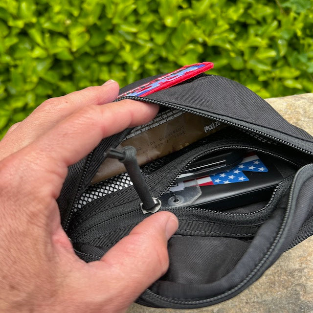 A hand opens the Tactical Fanny Pack - Cordura, revealing a pen with an American flag design and other small items inside its padded pocket.