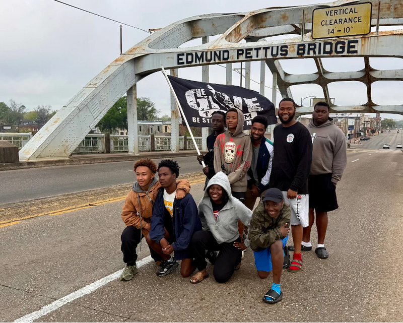 Eight young people pose and smile on the Edmund Pettus Bridge, holding a flag with text—celebrating camaraderie after participating in the Ruck Workout and Film Screening for The Give Team, inspired by GORUCK HQ in Jacksonville Beach, FL.