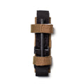 A Tourniquet Attachment in black, fastened with two tan hook-and-loop retention straps, is photographed on a white background from a top view.