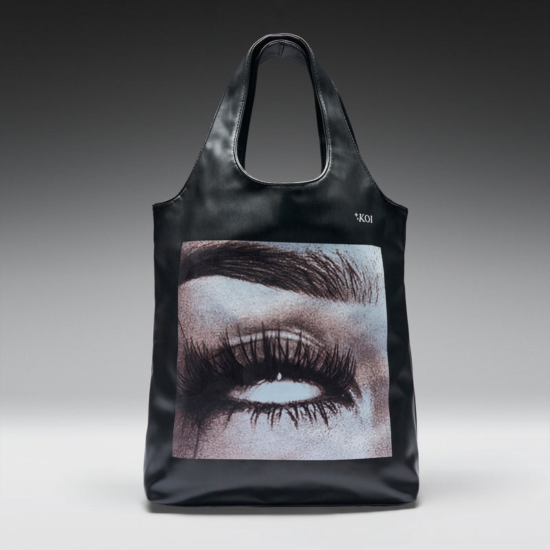 Black tote bag with eye design and 'Koi' branding on a gray background