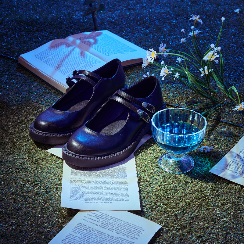 A pair of black faux leather Mary Jane shoes with safety pin buckles, displayed on a surface with a book, papers, and a glass of water.