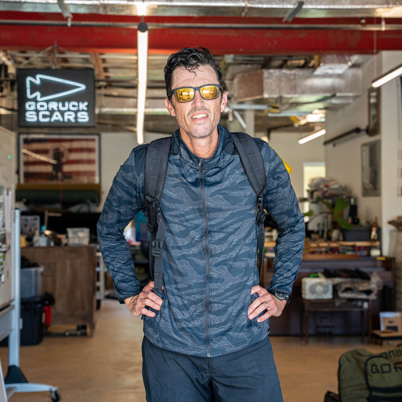 A man wearing sunglasses and a backpack stands in a workshop with a "GORUCK SCARS" sign in the background, dressed in the Warm Up Full Zip jacket featuring ToughDry shoulders.