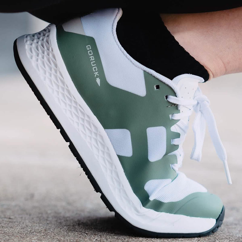 A close-up shot of a foot sporting the Women's Rough Runner - Seaspray by GORUCK, specifically designed for high mileage running. The shoe showcases a stylish green and white design with black laces and a textured white sole, set against concrete. Its Gradient Density EVA Midsole offers exceptional comfort and durability, making it perfect for conquering any Rough Runner challenge.