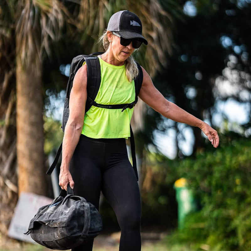 Woman wearing GORUCK gear, rucksack, and cap outdoors, carrying a training sandbag